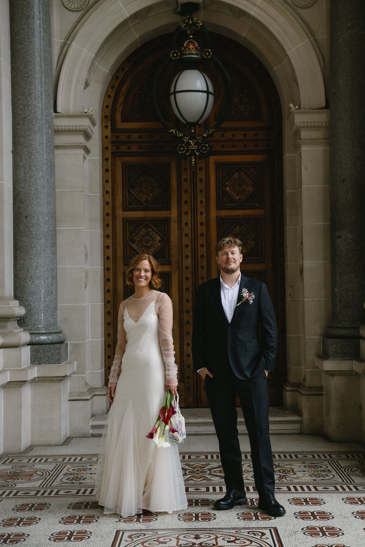 Bride and groom posing under ornate archway with decorative tiled floor and grand wooden doors.