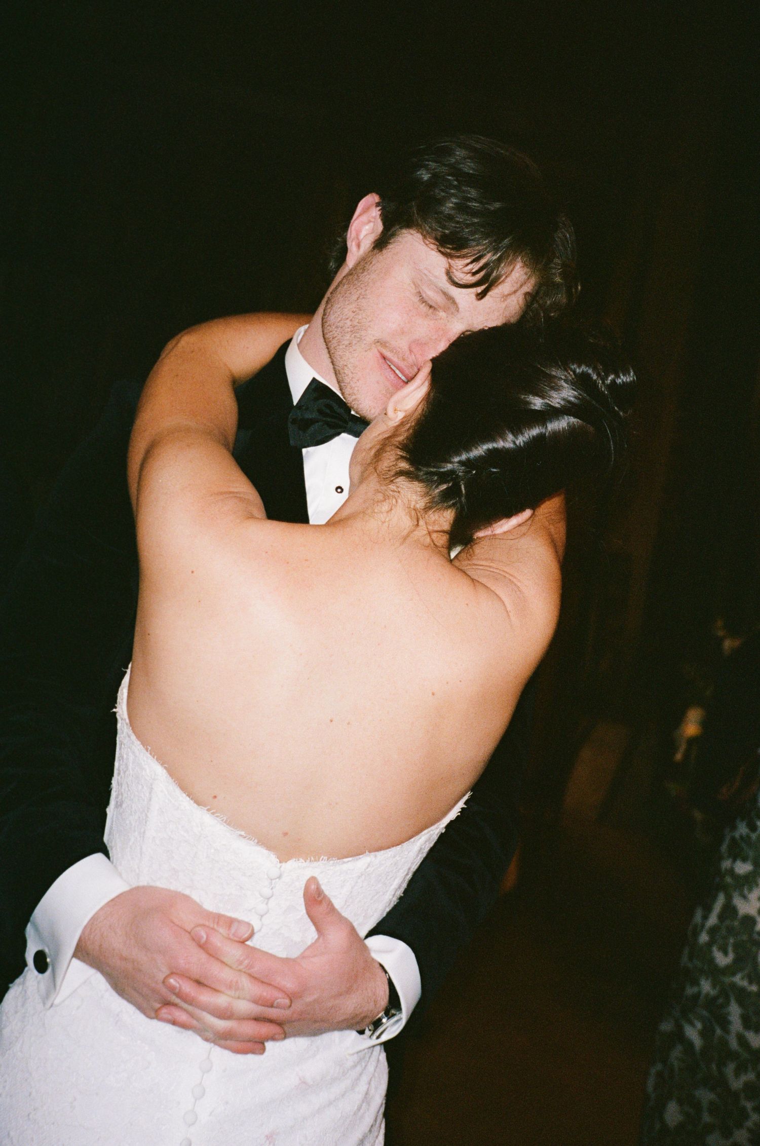 A bride and groom share an intimate first dance embrace, wearing a white backless gown and black tuxedo.