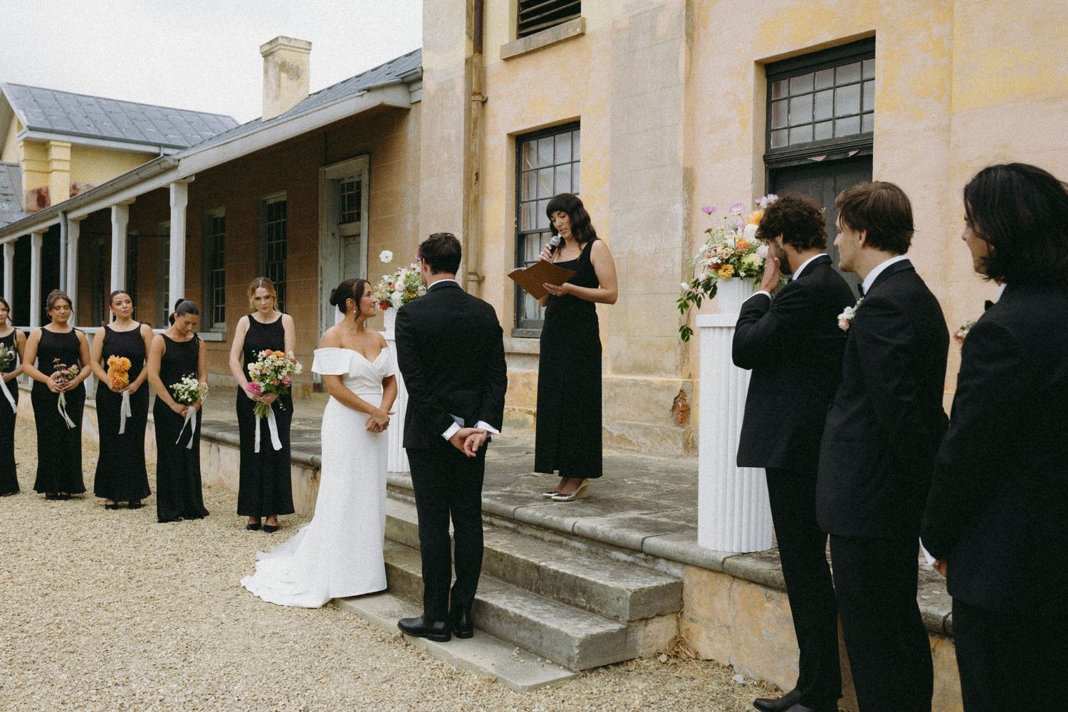 Elegant outdoor wedding ceremony with bridesmaids in black gowns and groomsmen in tuxedos at a historic stone venue.