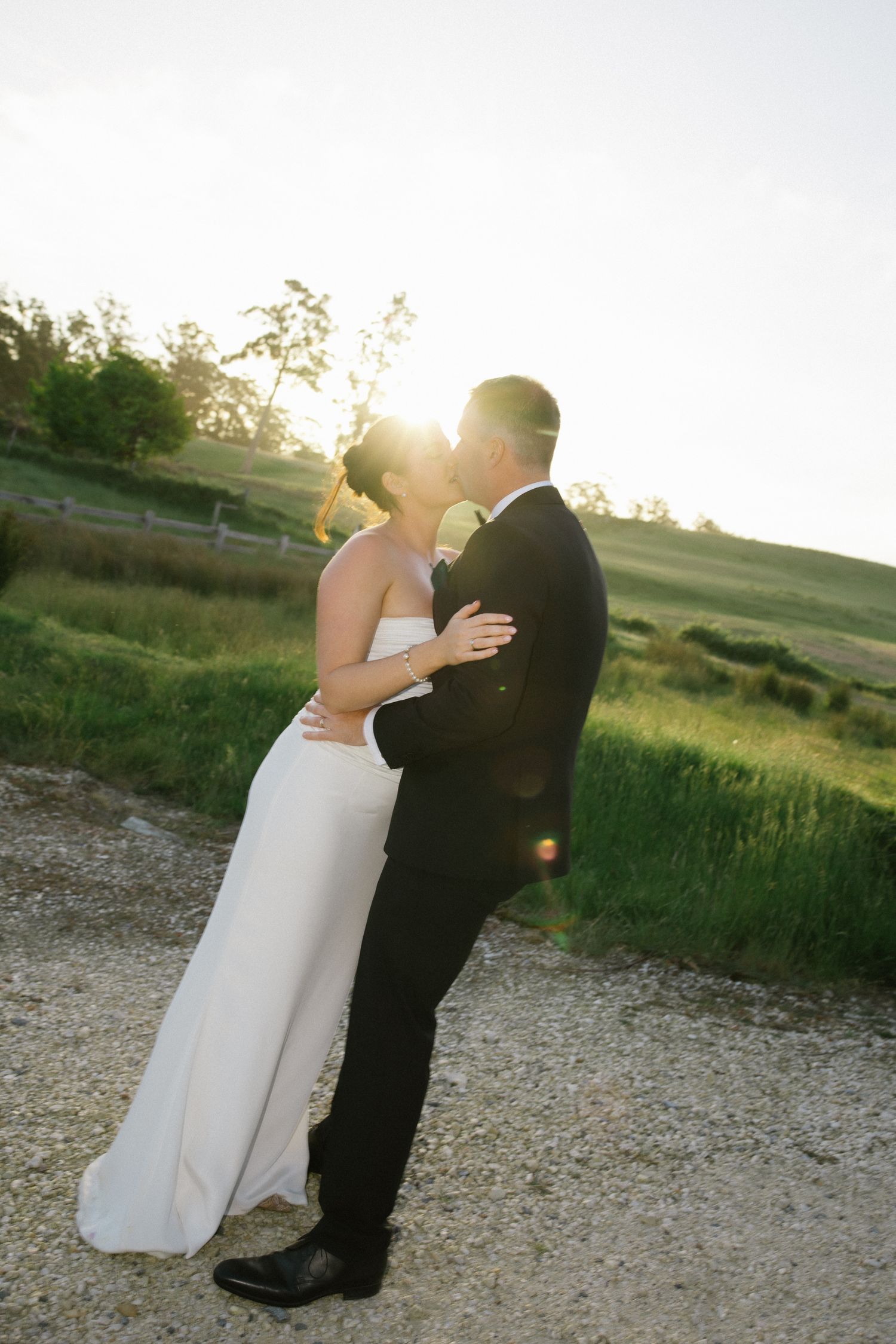 Bride and groom sharing a golden hour kiss on a gravel path surrounded by green fields.