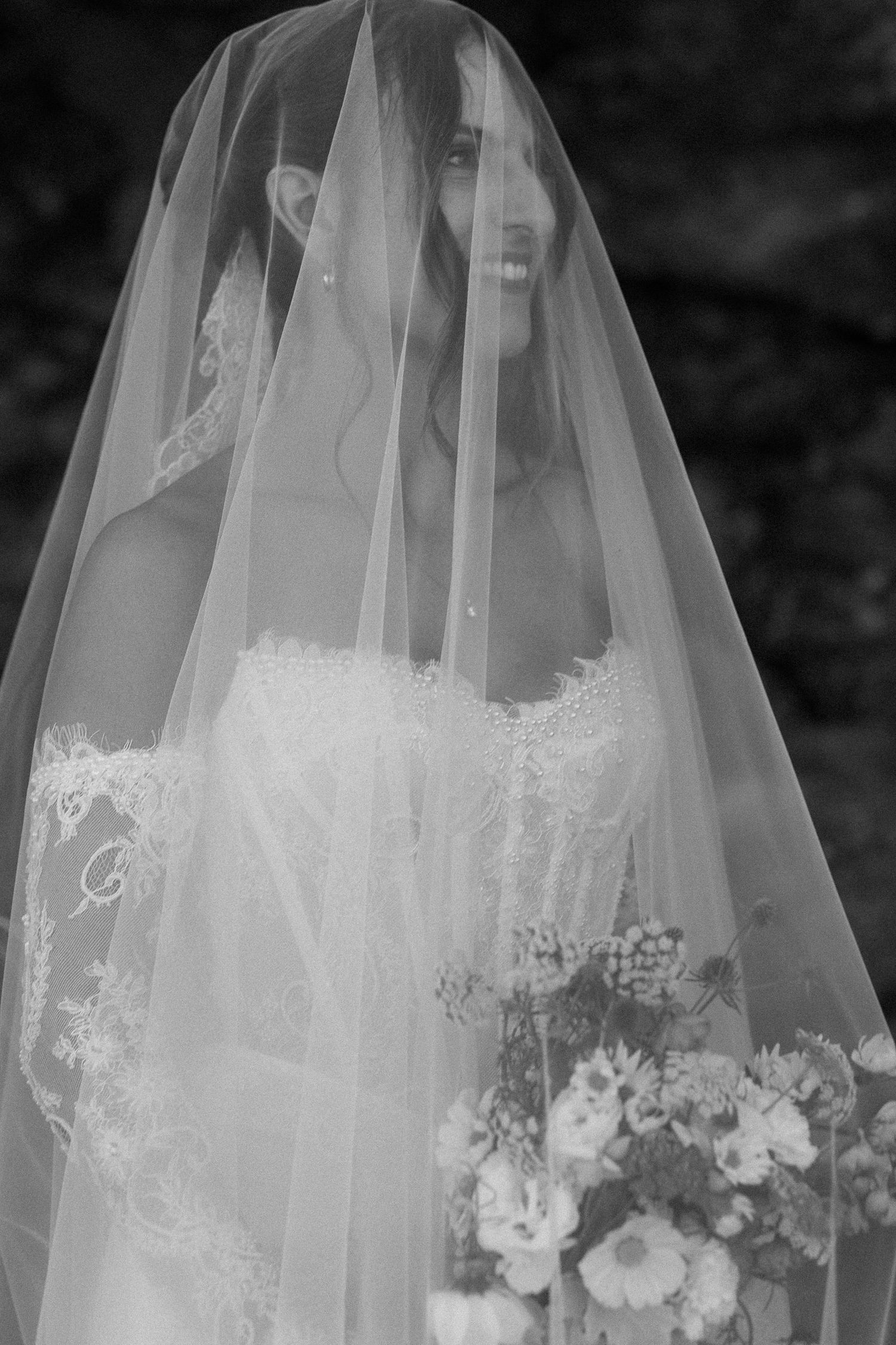 Black and white portrait of smiling bride draped in lace-edged veil holding a floral bouquet.
