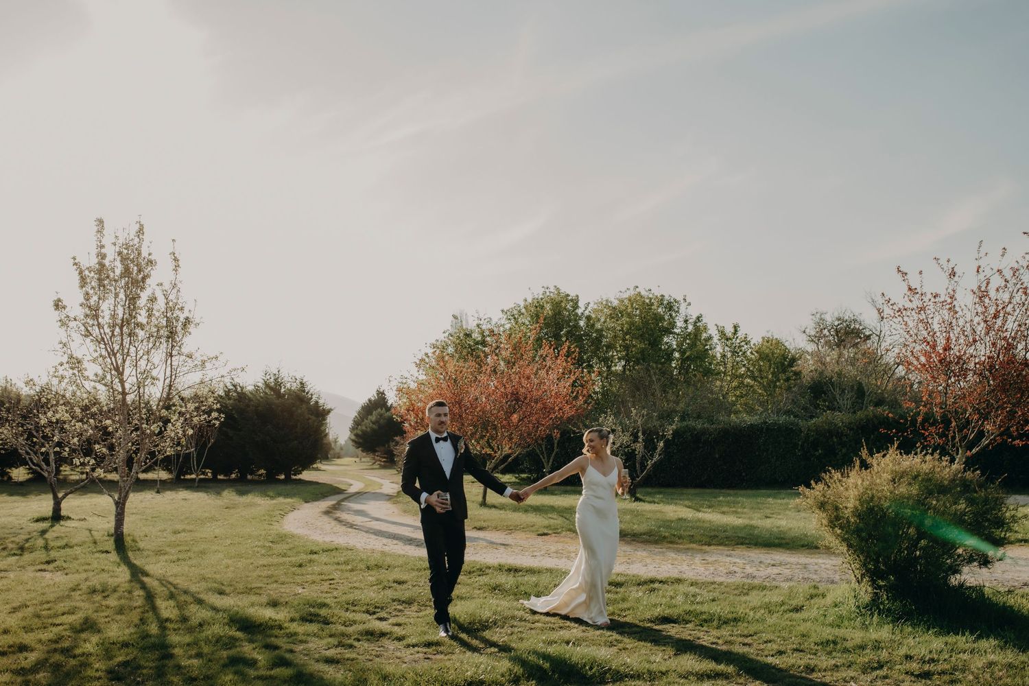 Bride and groom walk hand in hand through orchard garden on sunny wedding day.