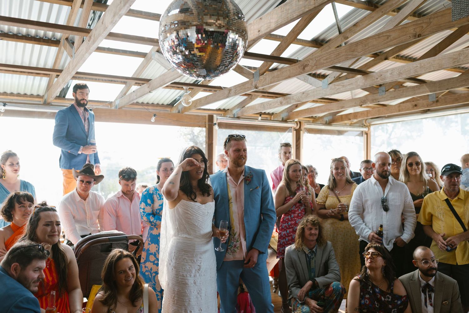 Couple exchanges vows at boho wedding ceremony under disco ball with guests seated in rustic outdoor venue.