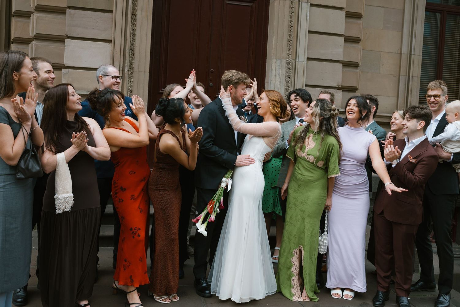 Joyful newlyweds kiss surrounded by cheering wedding guests outside grand stone building entrance.