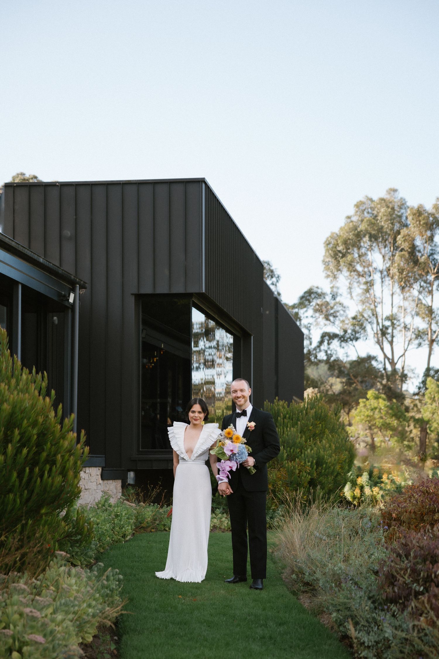 Bride and groom standing outside modern black building surrounded by lush gardens.