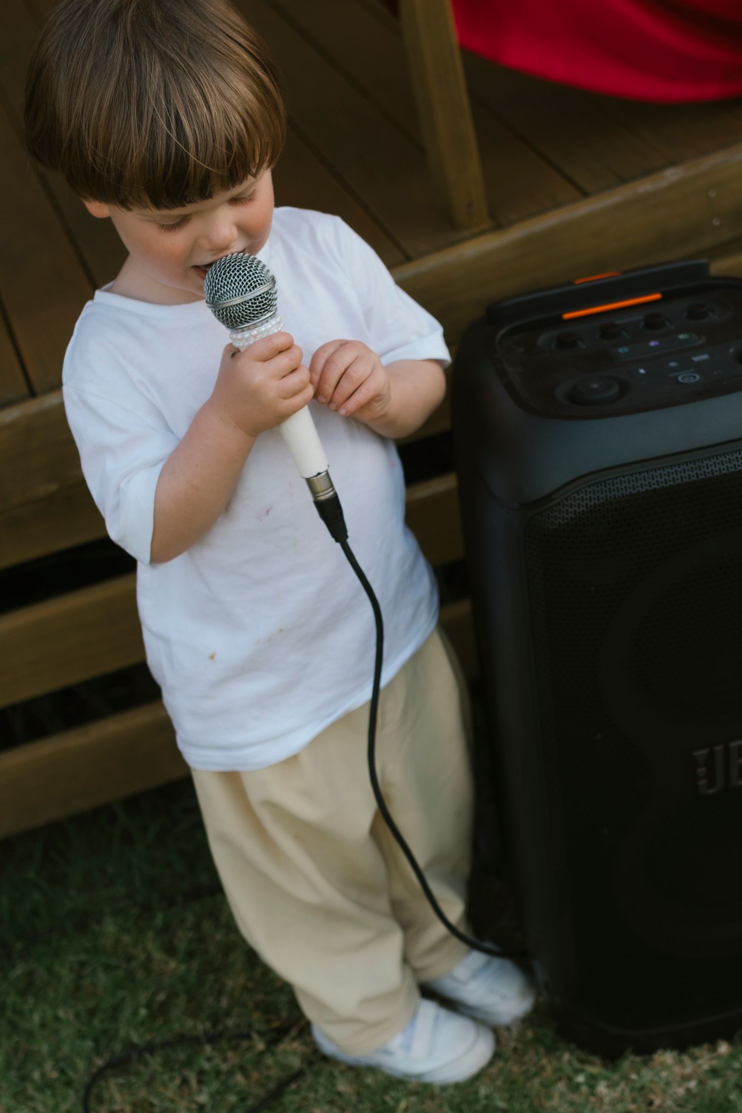 Young boy in white t-shirt singing into a wired microphone outdoors next to a black JBL speaker.