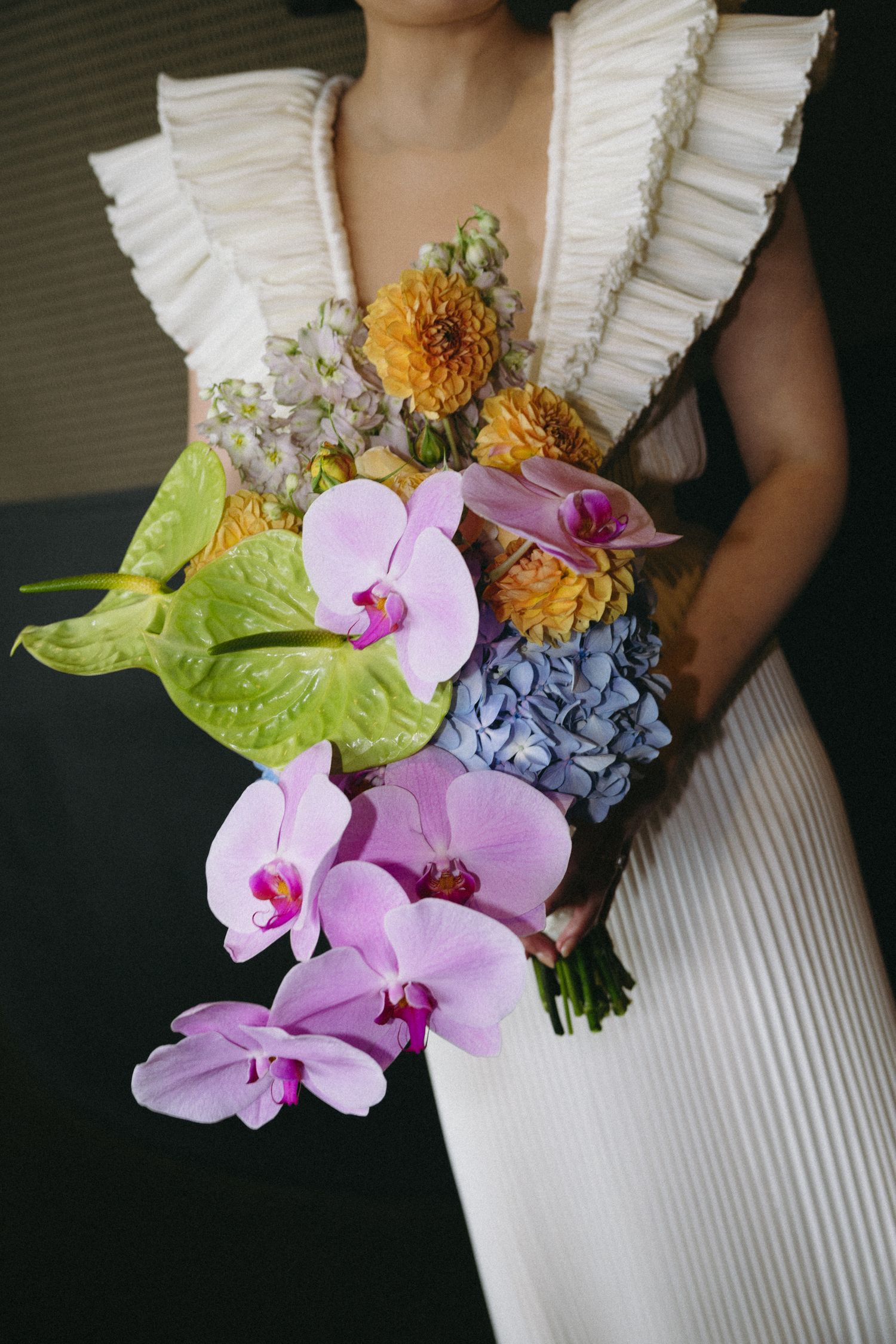 Bride in white ruffled gown holding colorful bouquet with pink orchids, blue hydrangeas, and yellow dahlias.