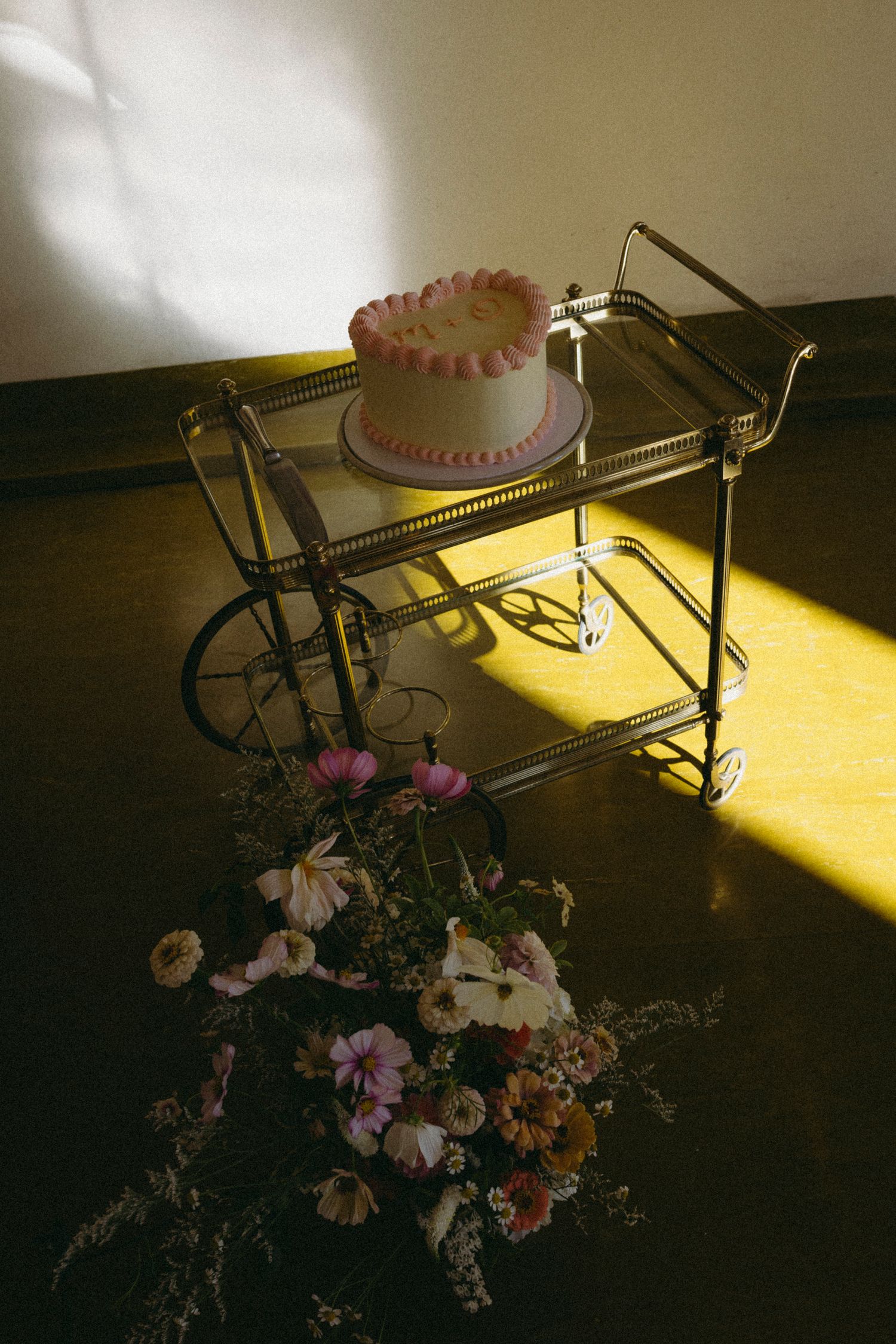 Single-tier white wedding cake with pink ruffled border displayed on vintage gold bar cart with flowers.