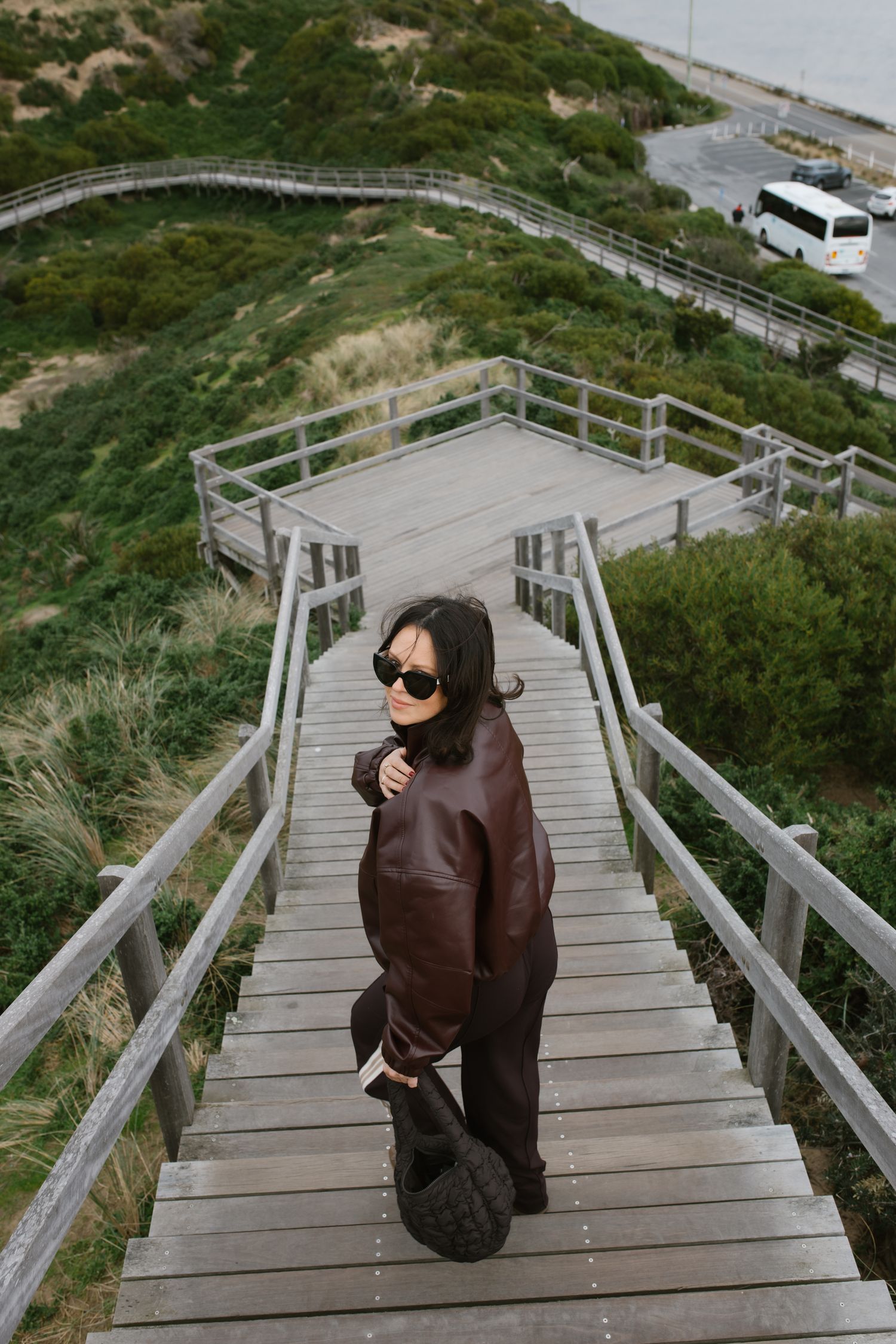 Woman in brown leather jacket and sunglasses walks up wooden coastal boardwalk stairs.