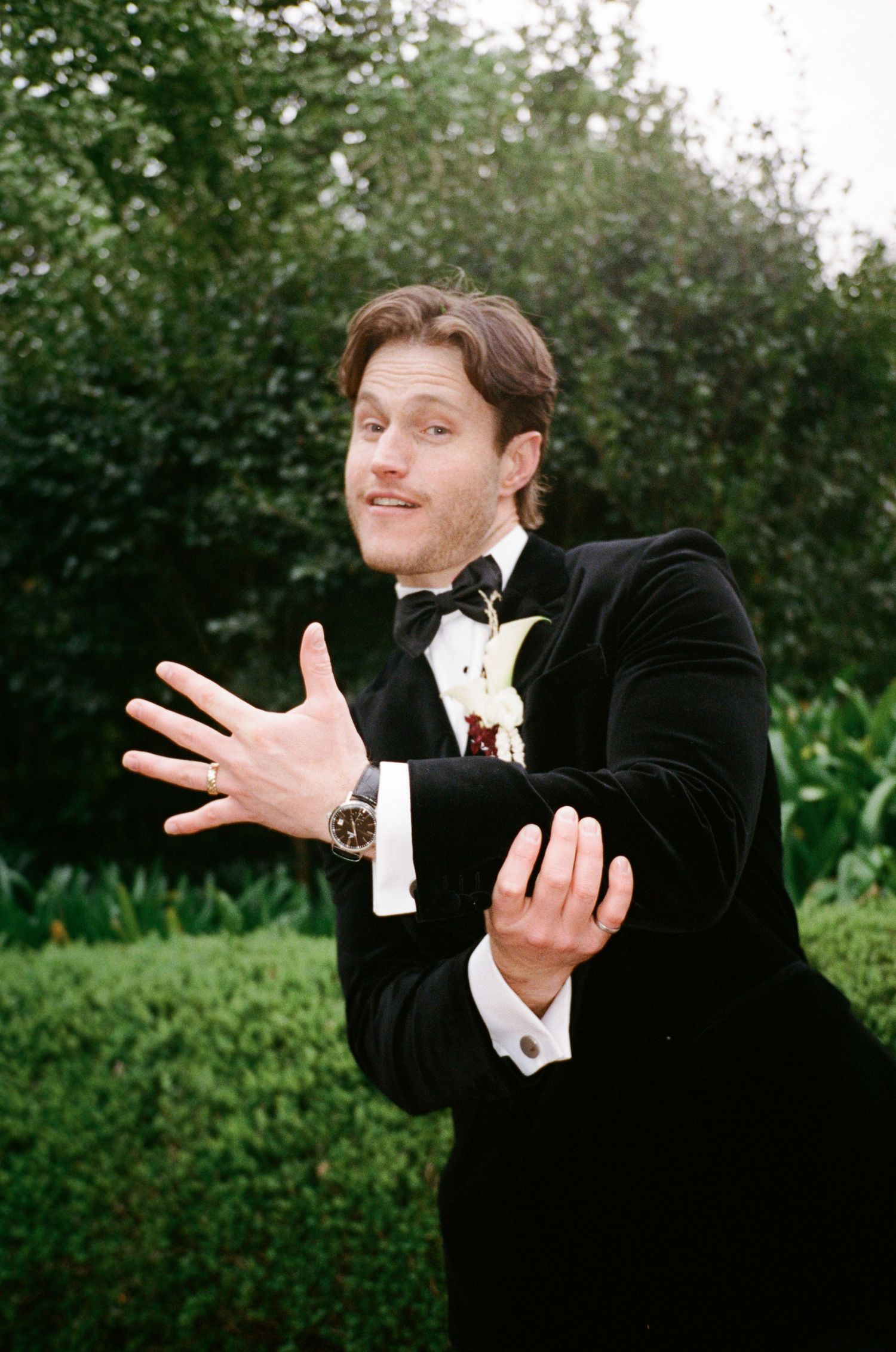 Man in black velvet tuxedo with bow tie and boutonniere gestures expressively outdoors among lush green hedges.
