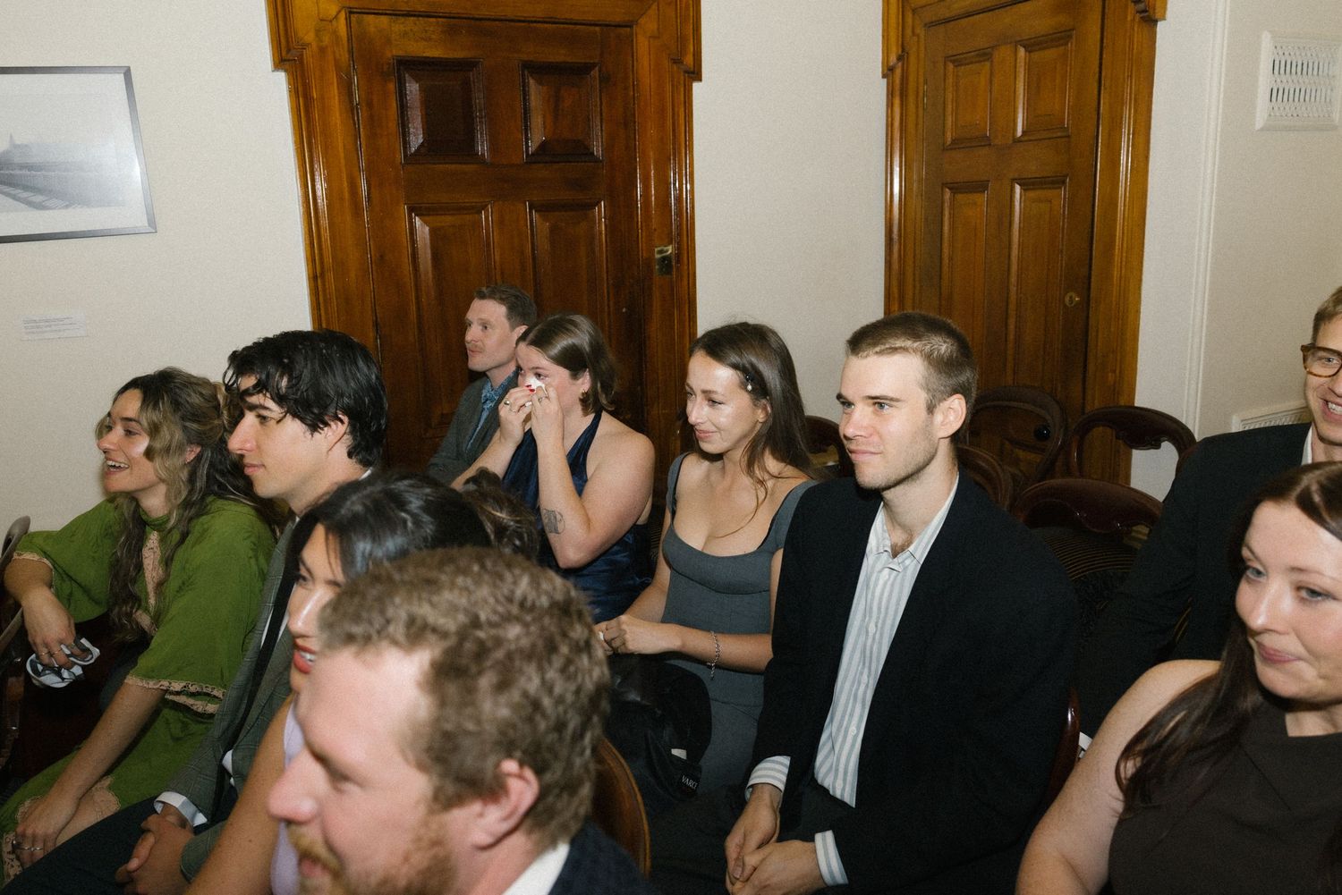 Wedding guests seated in rows inside ornate room watching civil marriage ceremony attentively.