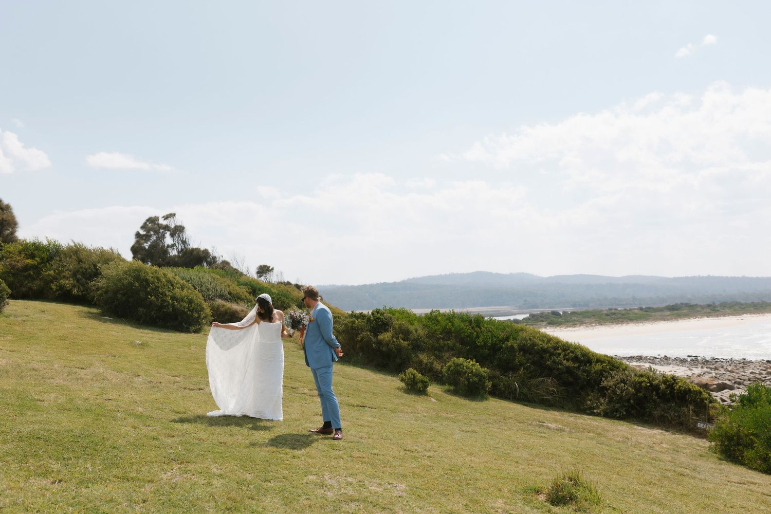 A couple walks hand-in-hand on a grassy hillside overlooking a scenic coastal estuary on their wedding day.