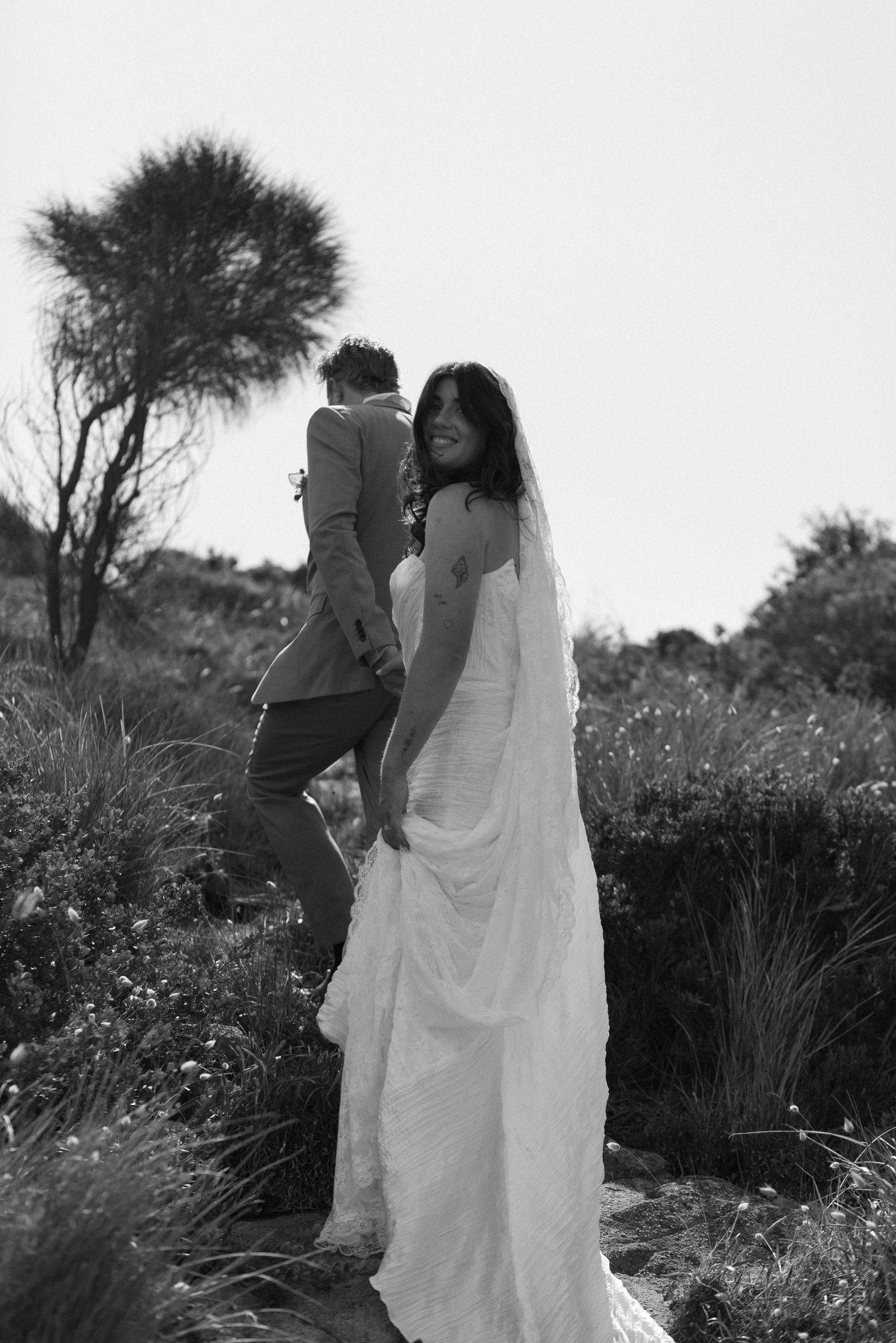 Black and white photo of smiling bride in lace gown holding grooms hand walking through nature.