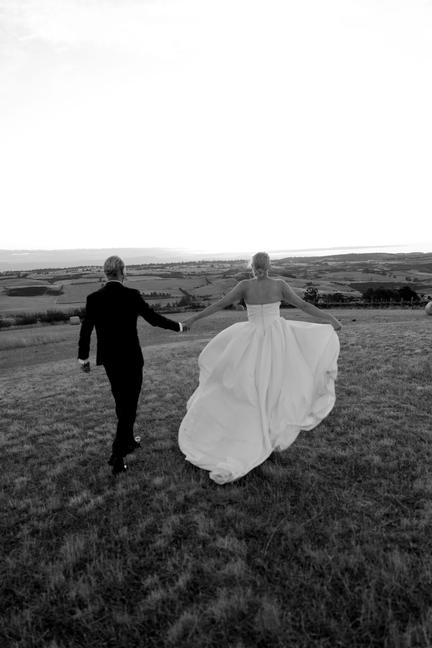 Bride and groom holding hands running through open fields in a black and white wedding photo.