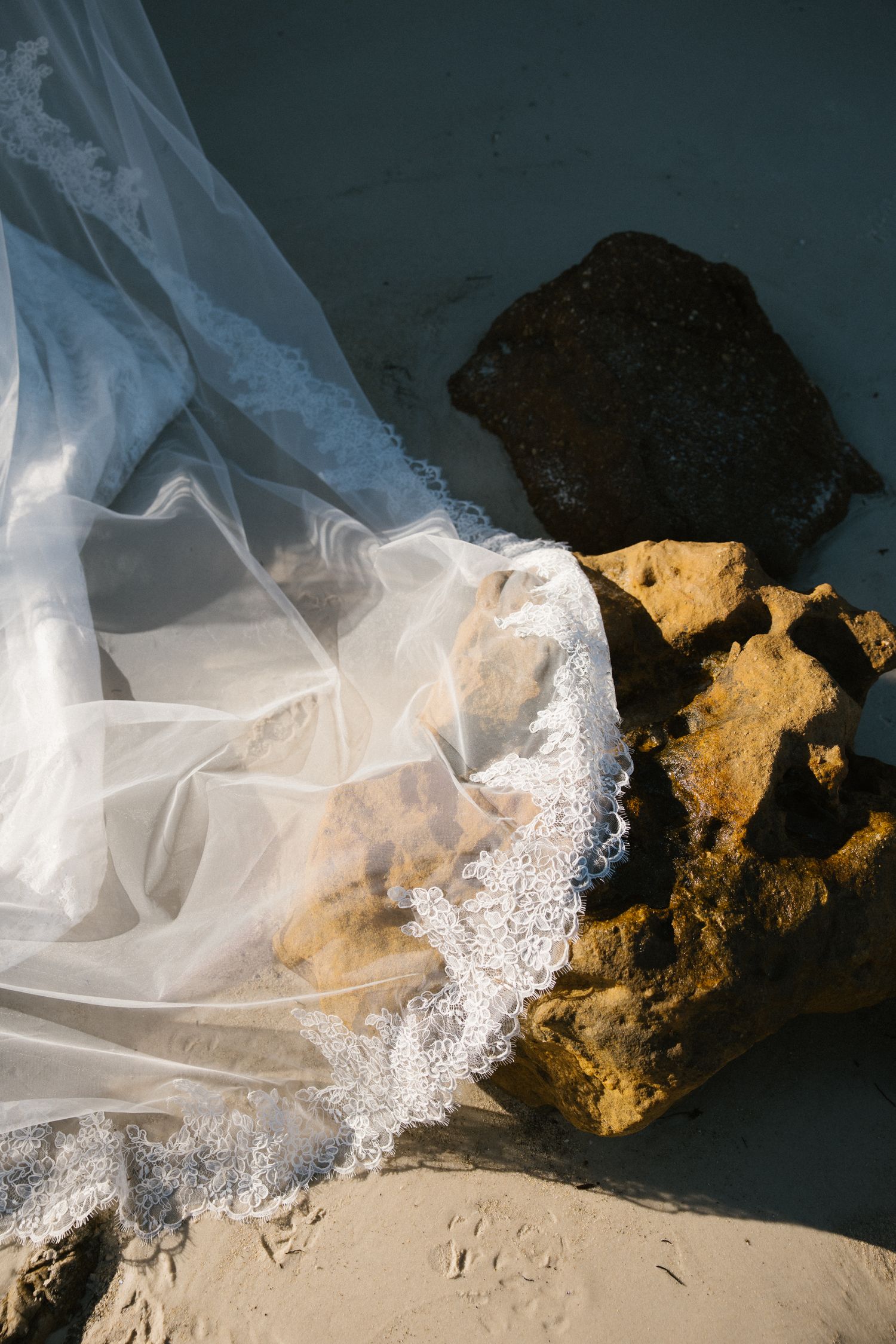 Delicate lace-trimmed wedding veil draped over sandy beach rocks in natural sunlight.