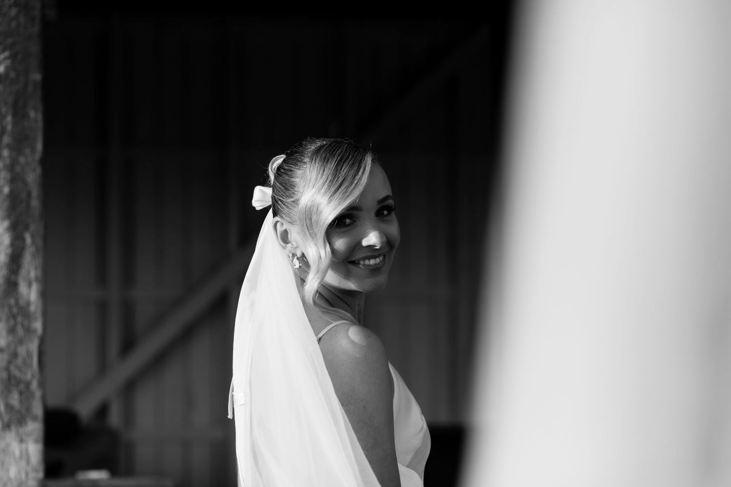 Black and white portrait of a smiling bride in a veil looking over her shoulder in a barn doorway.