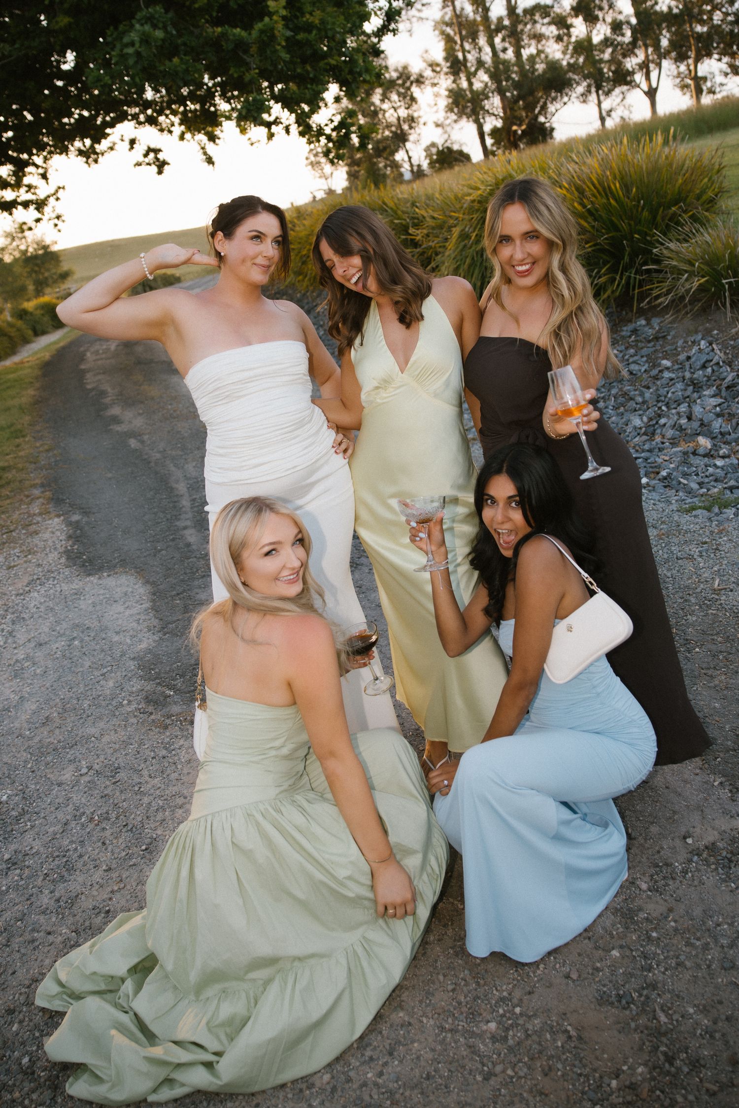 Group of five stylish wedding guests posing on a gravel path at sunset with wine glasses.