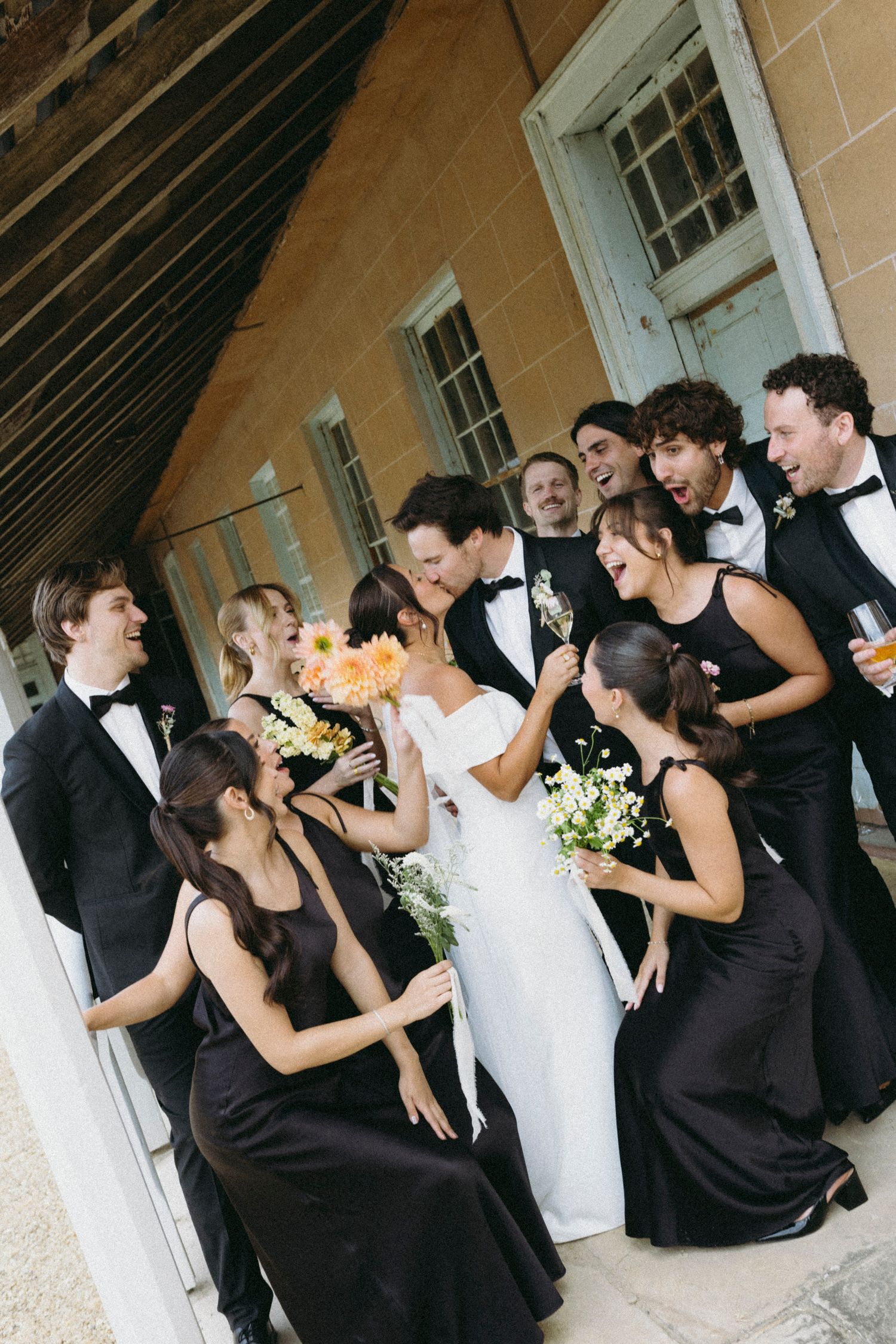 Laughing bride and groom celebrate with wedding party dressed in black tuxedos and gowns outdoors.