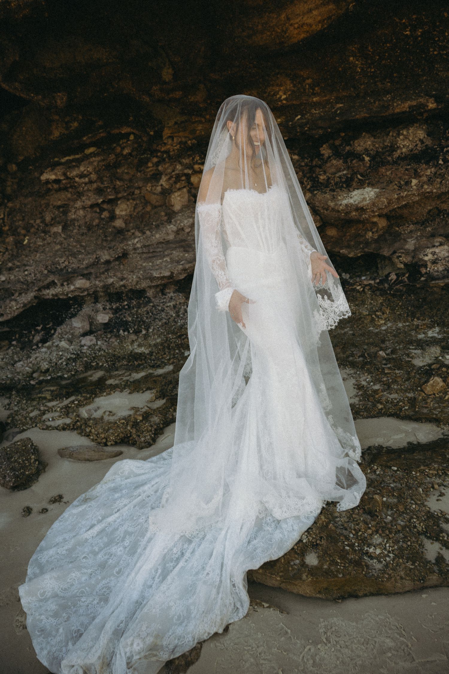 Bride in fitted lace wedding gown with long cathedral veil posing against dramatic rocky backdrop.