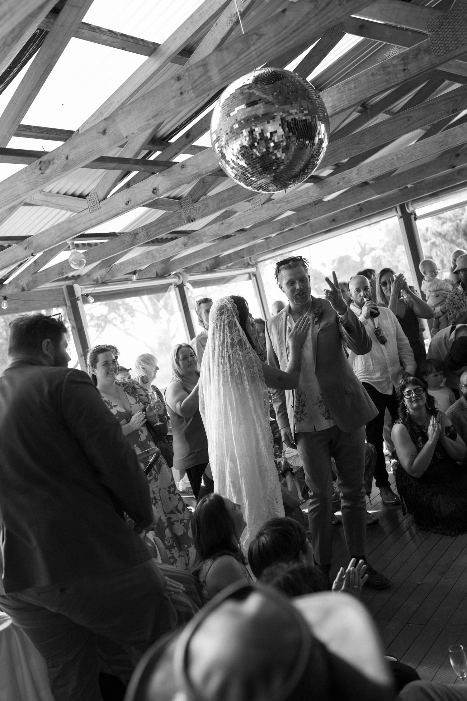 Black and white photo of a bride and groom sharing first dance under a disco ball at a rustic wedding venue.