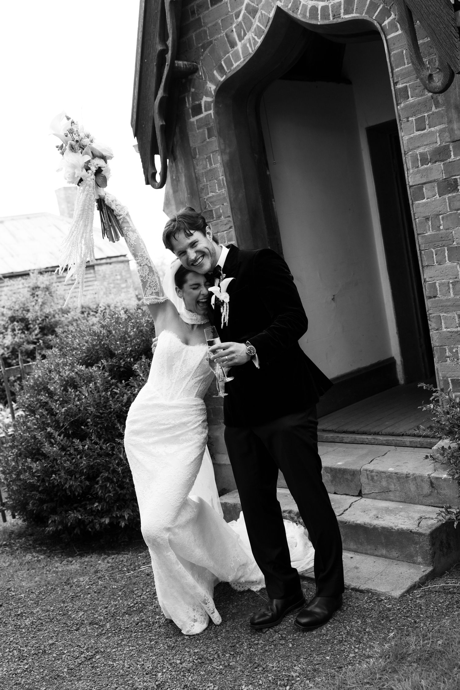 Joyful bride and groom celebrate outside a brick church with champagne and bouquet.