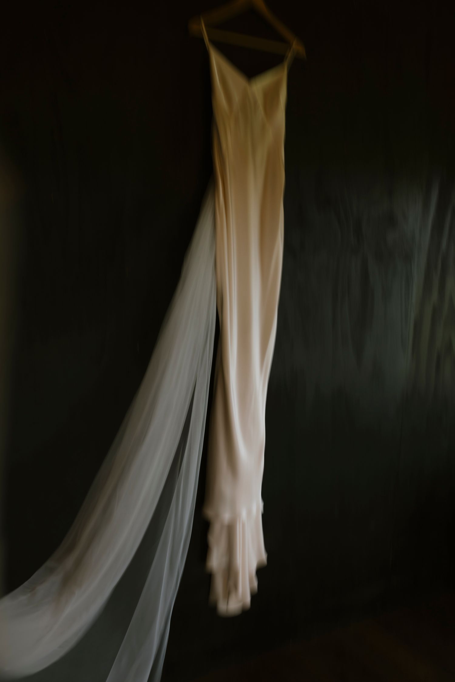 Elegant white wedding dress with veil hanging on wooden hanger against dark wall.