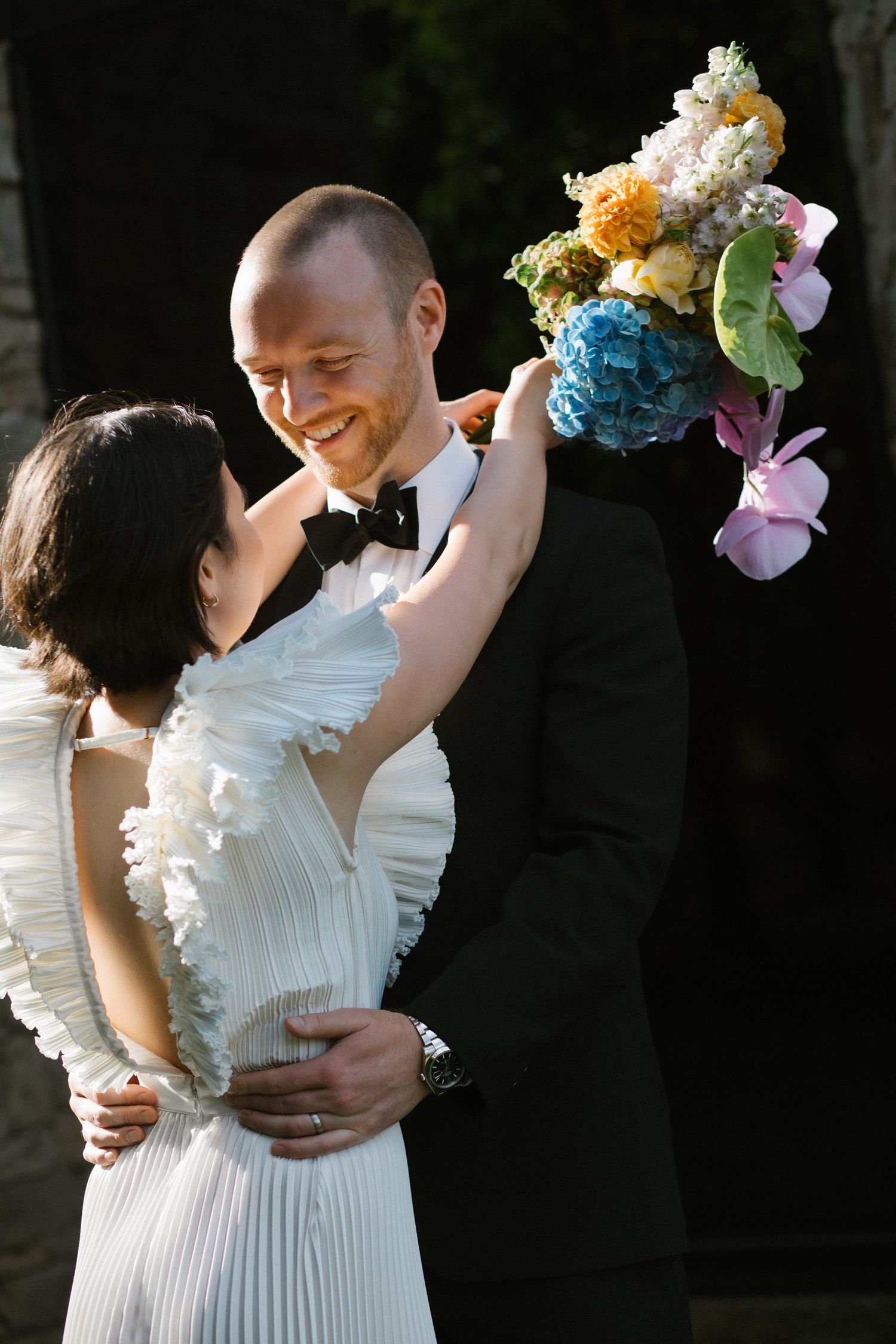 Joyful bride raising colorful bouquet while embracing groom in black tuxedo at outdoor wedding.