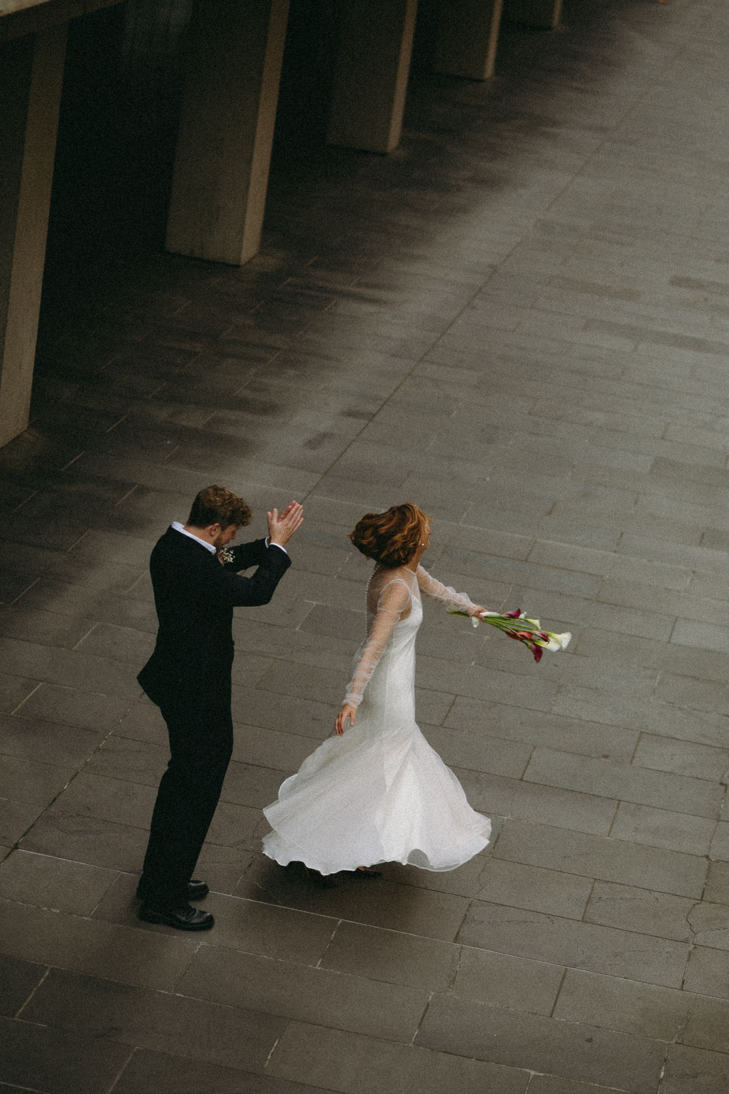Aerial view of bride and groom dancing on stone plaza, bride twirling in flowing white gown with bouquet.