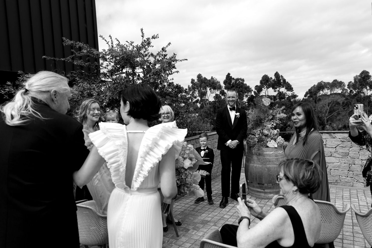 Black and white photo of bride approaching groom and wedding guests at outdoor ceremony venue.