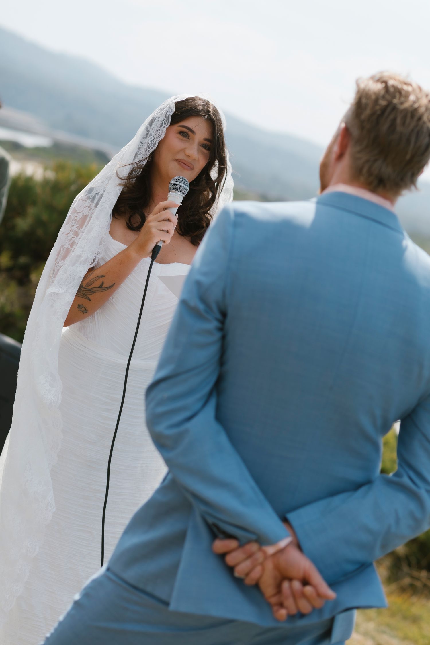 Bride in white gown and veil recites vows into microphone to groom in blue suit outdoors.