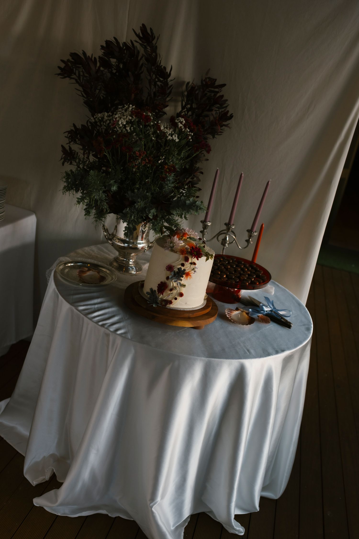 Elegant wedding cake table with white tablecloth, floral arrangement, red candles, and decorated cake in a tent setting.