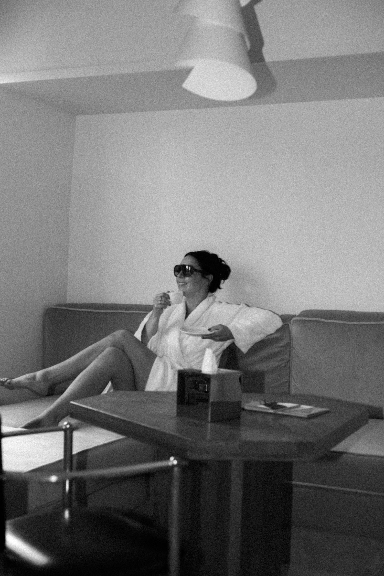 Black and white photo of a bride in a white robe and sunglasses relaxing on a sofa with coffee.