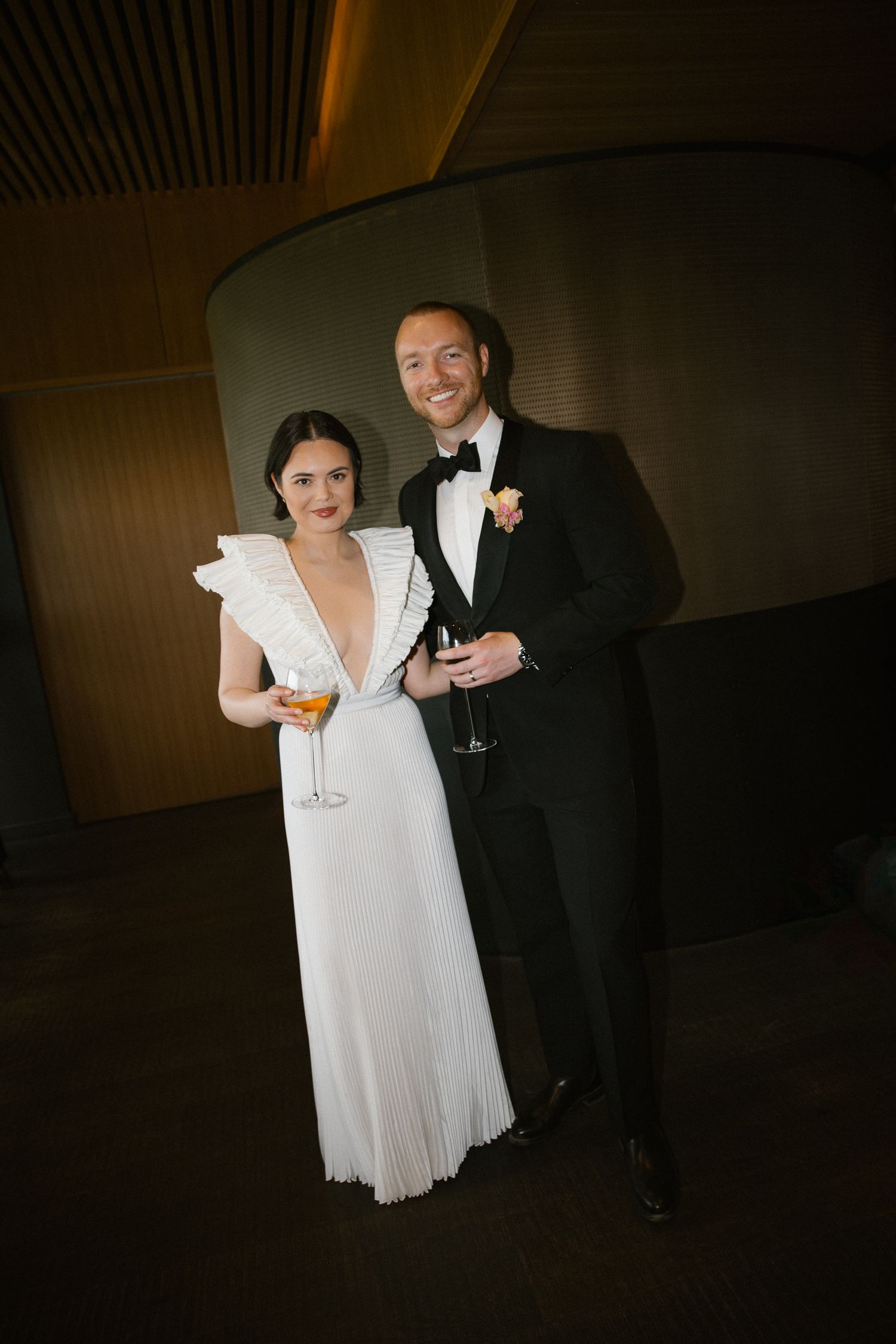 Smiling bride and groom in white gown and black tuxedo holding cocktails at elegant wedding reception.