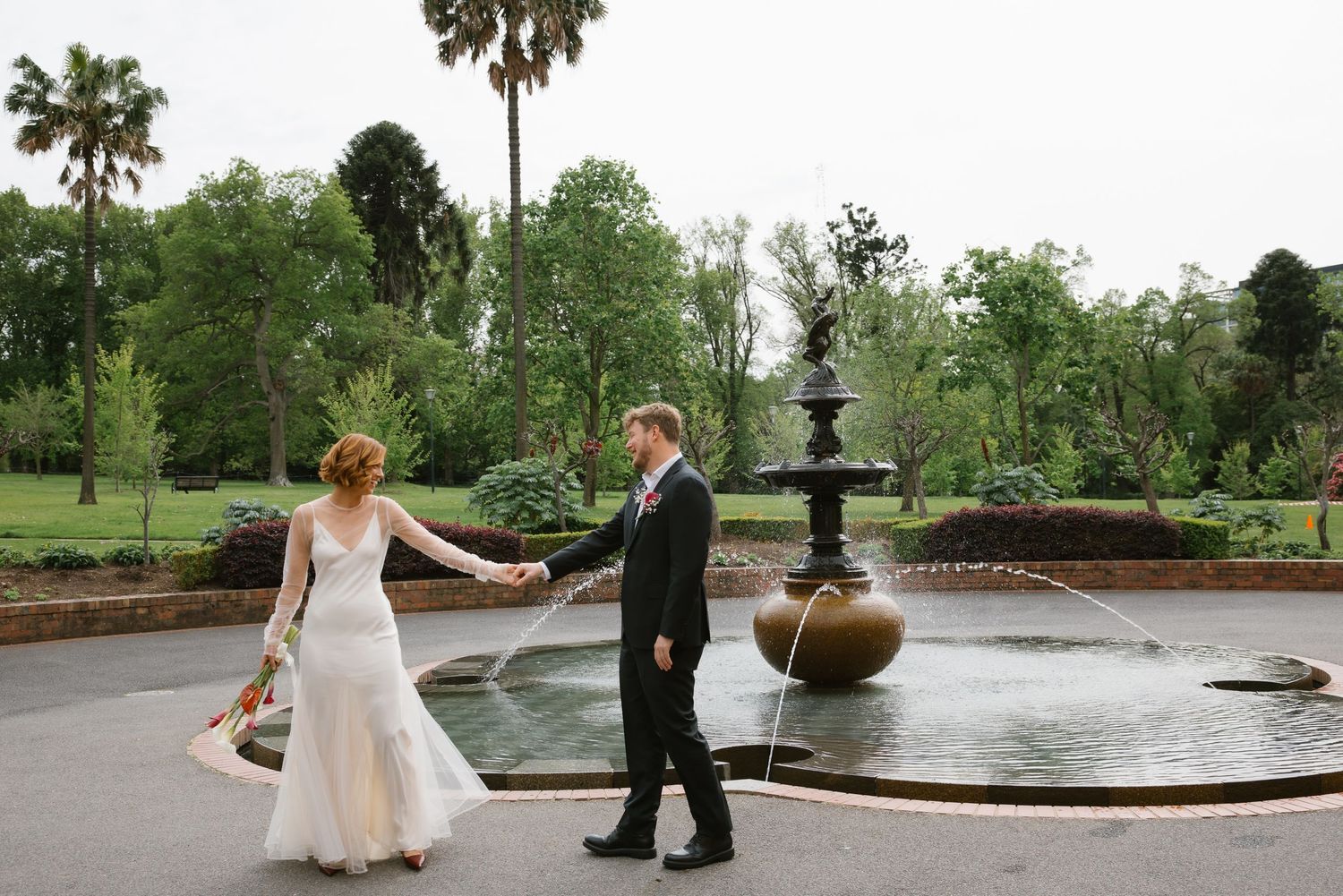 Bride and groom walking hand in hand past an ornate fountain in a lush garden park.