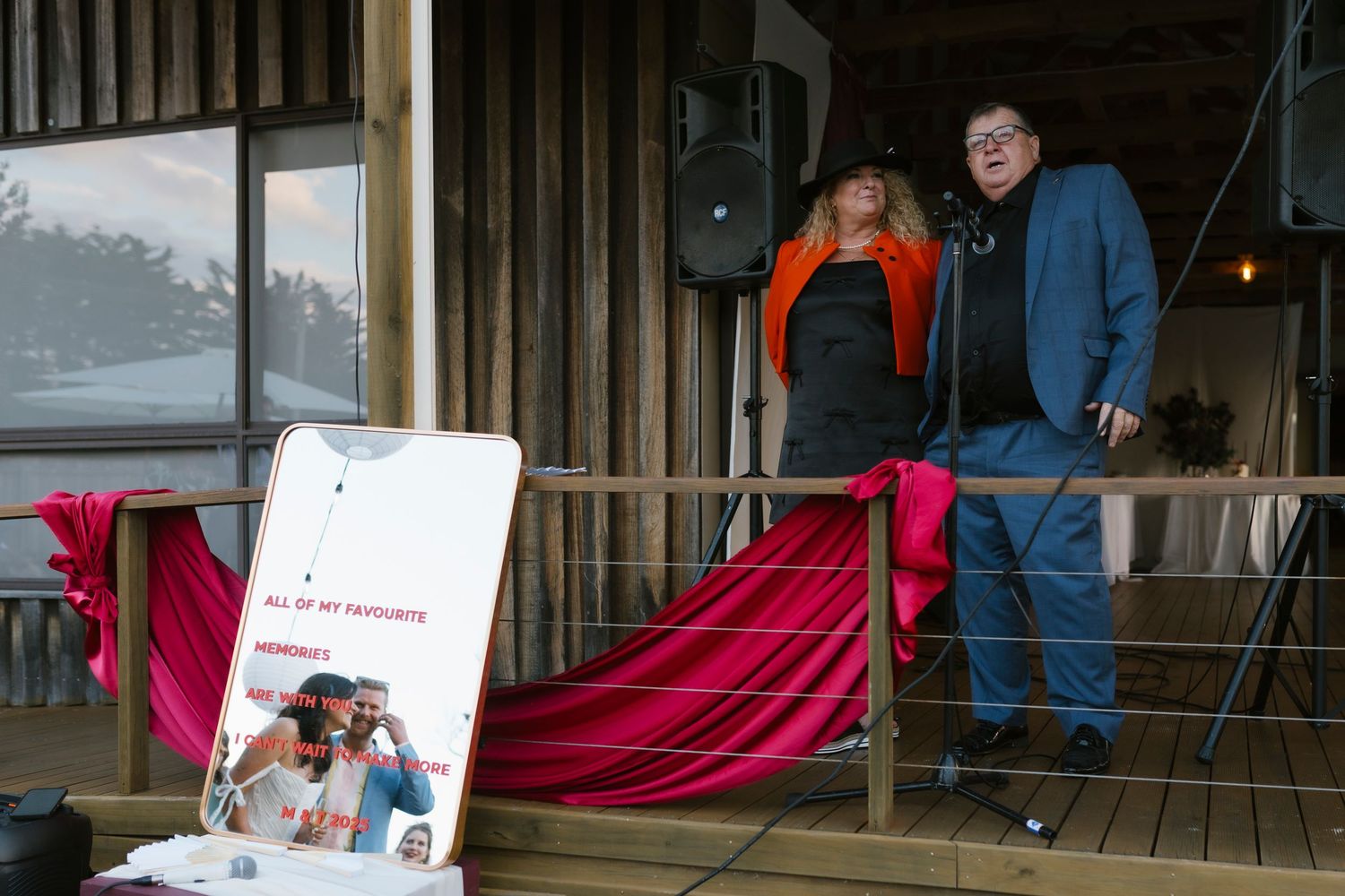 Two people stand on a wooden deck near a speaker and red drape at an outdoor event.