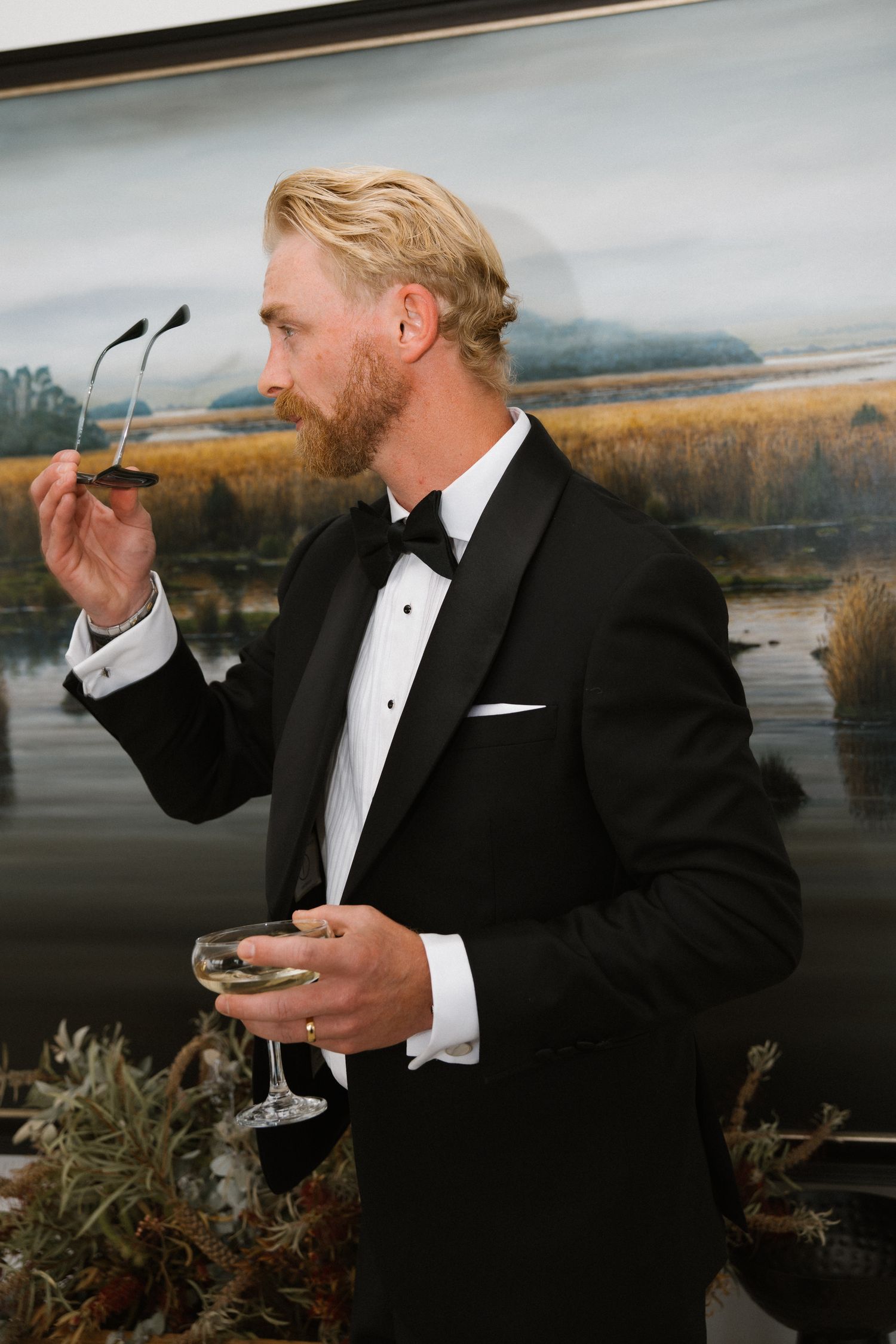 Elegant groom in black tuxedo holding champagne glass and sunglasses beside landscape painting.