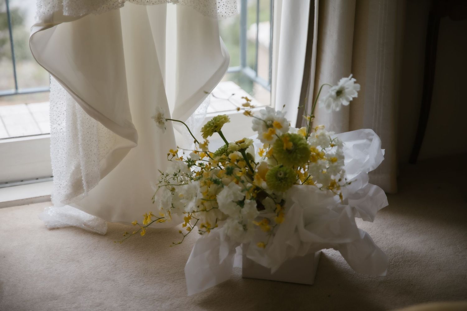 Delicate bridal bouquet of white and yellow wildflowers with tulle ribbon beside wedding veil at window.
