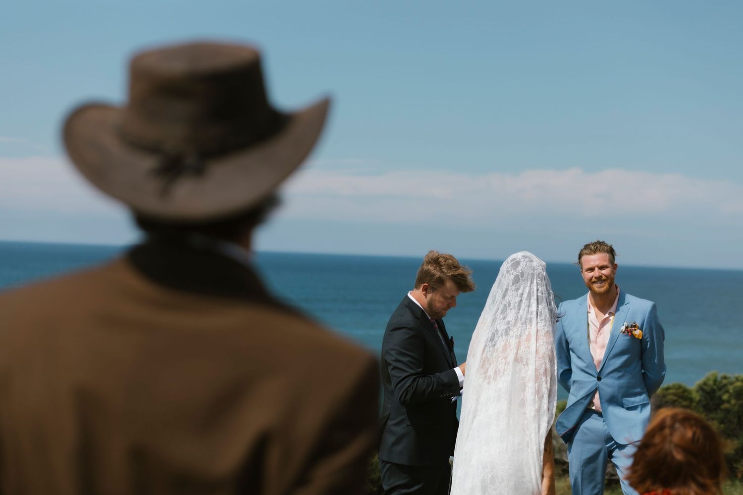 Outdoor beach wedding ceremony with groom in blue suit, bride in veil, officiant, and guest in cowboy hat.