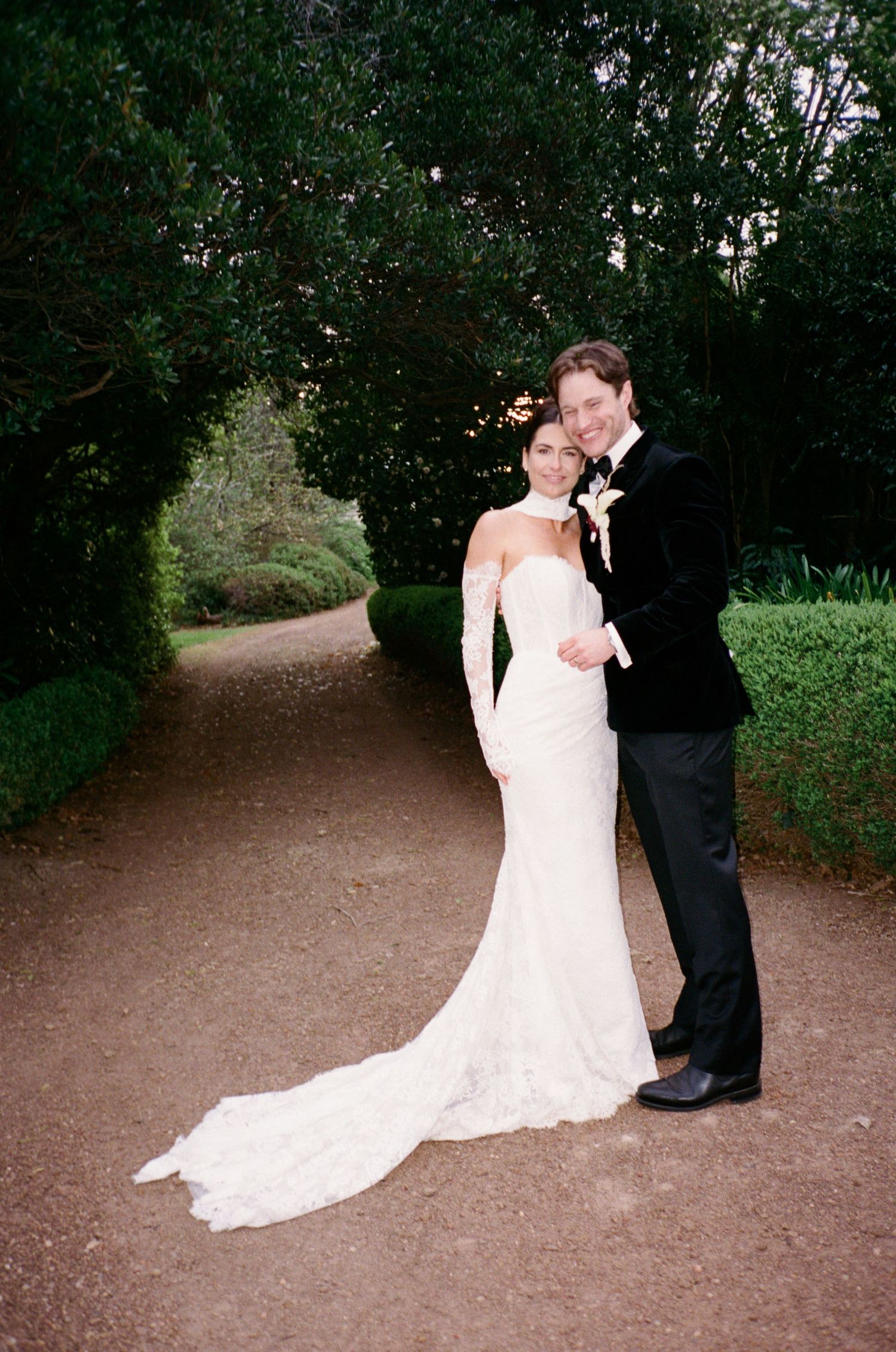 Bride in white lace gown with train and groom in black tuxedo posing on garden path surrounded by lush greenery.