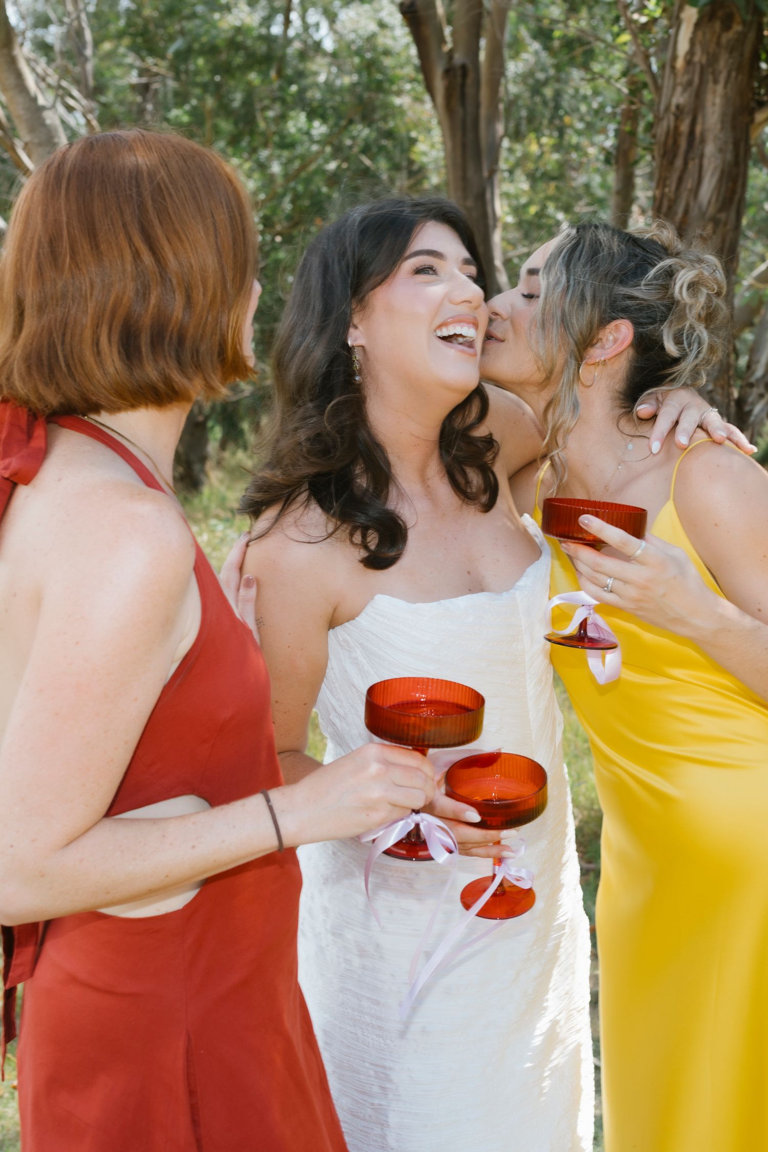Bride in white dress laughing with bridesmaids in red and yellow holding ruby red wine glasses outdoors.