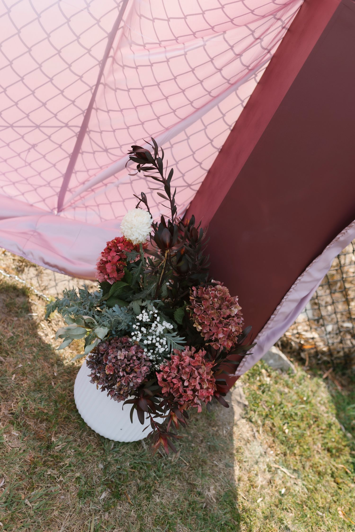 Burgundy and pink wedding arch decorated with dried hydrangeas, greenery, and white flowers on grass.