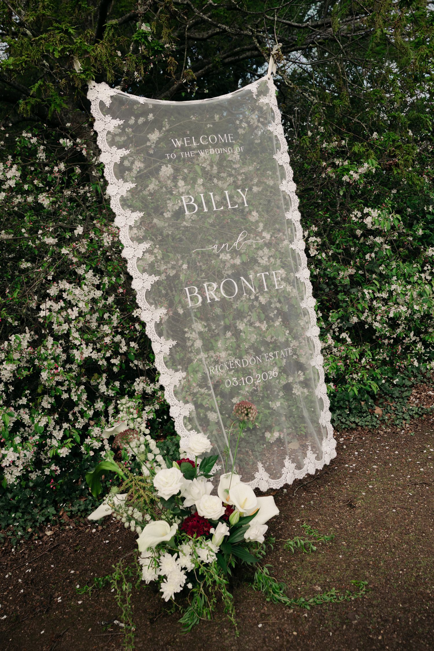 Elegant lace wedding welcome sign for Billy and Bronte displayed among white flowering garden shrubs.