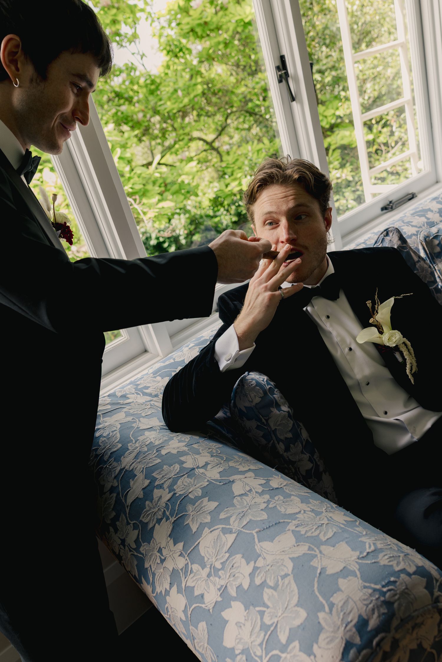Two grooms in tuxedos share a joyful moment on a floral sofa near a sunlit window.