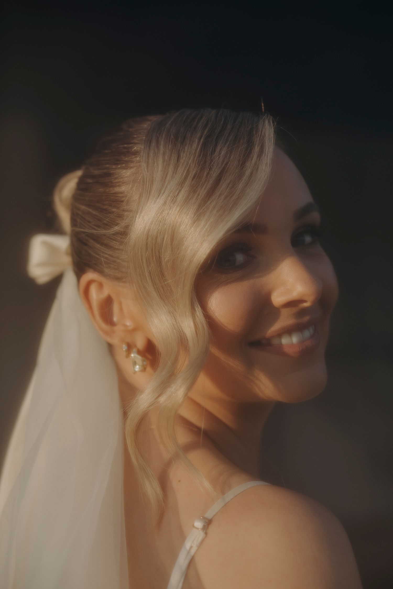 Beautiful blonde bride smiling in golden sunlight wearing a white bow, veil, and pearl earrings.