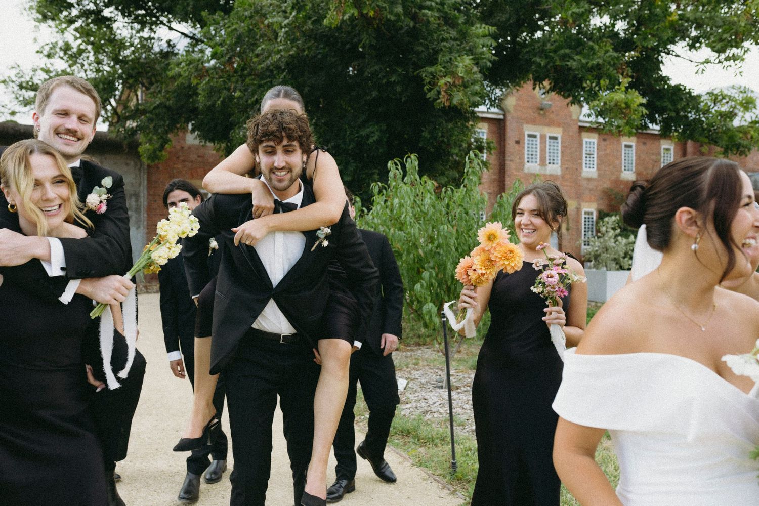 Fun wedding party moment with groomsman giving piggyback ride outside historic brick building.