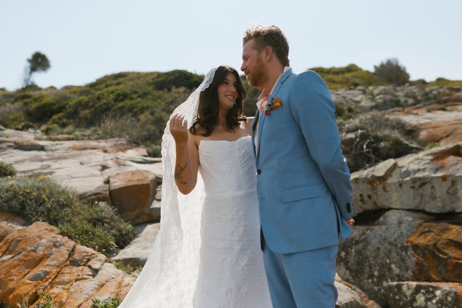 A bride in a white gown and veil stands with a groom in a light blue suit on rocky coastal terrain.
