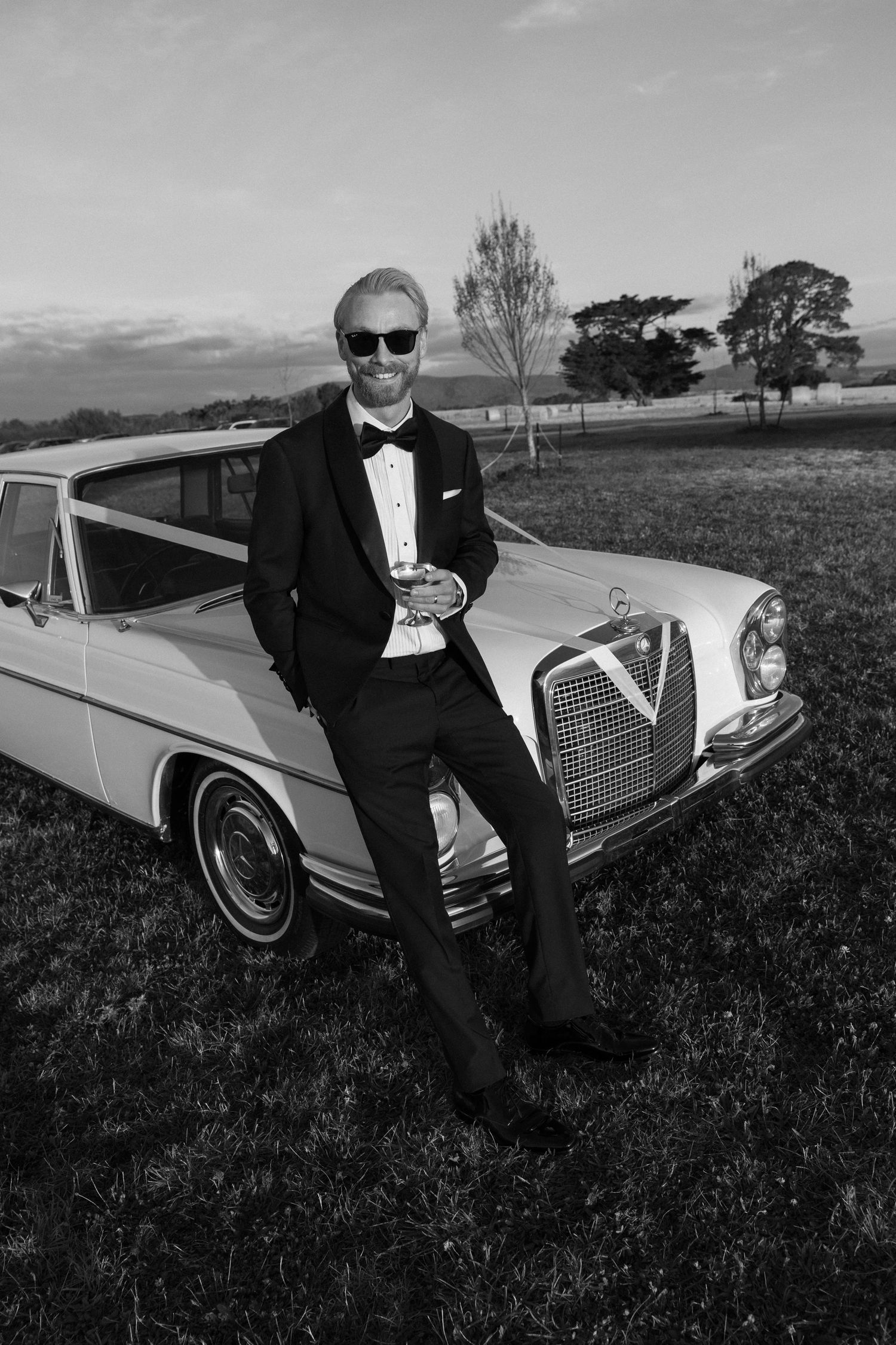 Groom in tuxedo and sunglasses leaning against vintage Mercedes in black and white portrait.