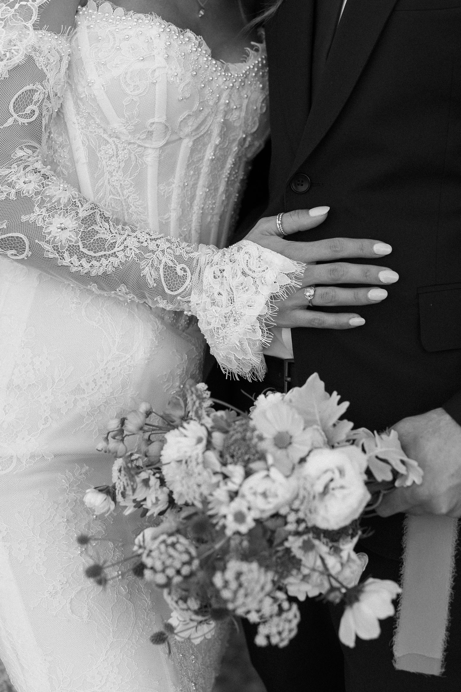 Black and white close-up of bride in lace gown holding bouquet with groom in black suit.