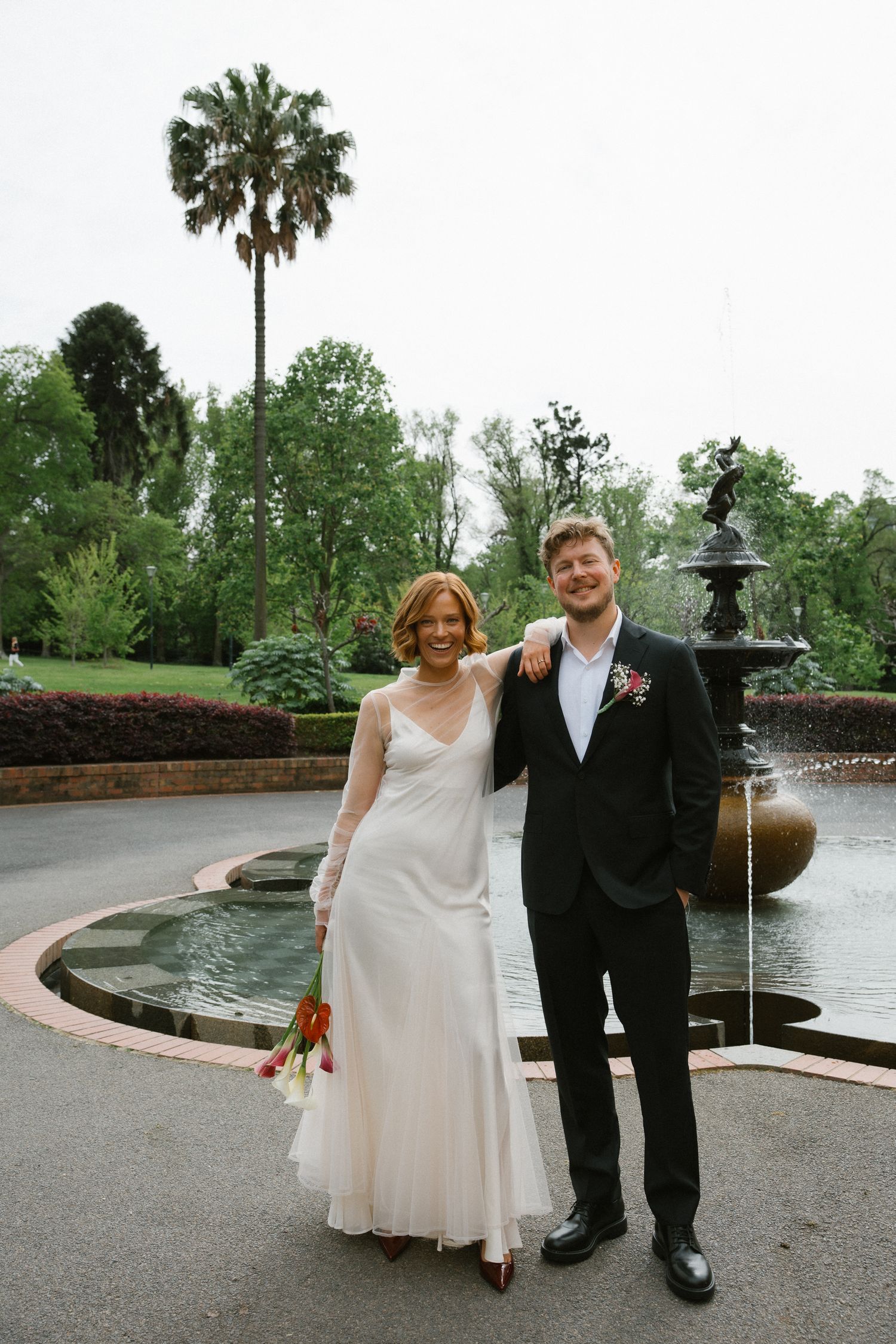 Smiling bride and groom pose together in front of ornate fountain in lush garden setting.