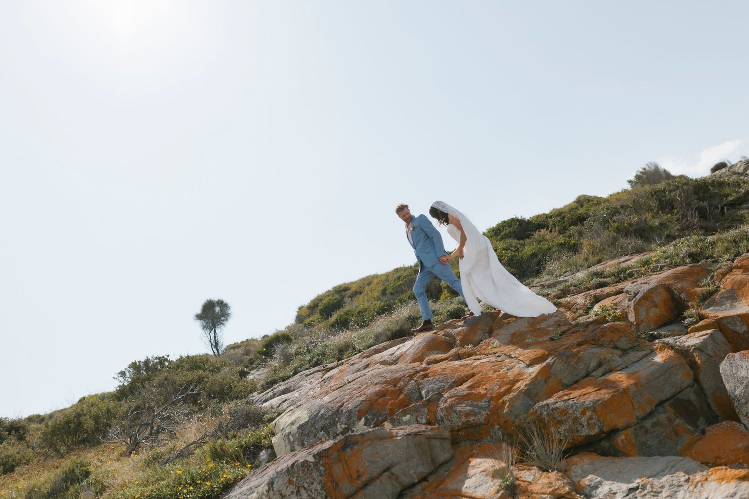 Couple in wedding attire climbing rocky coastal hillside, bride in white gown, groom in blue suit, sunny day.